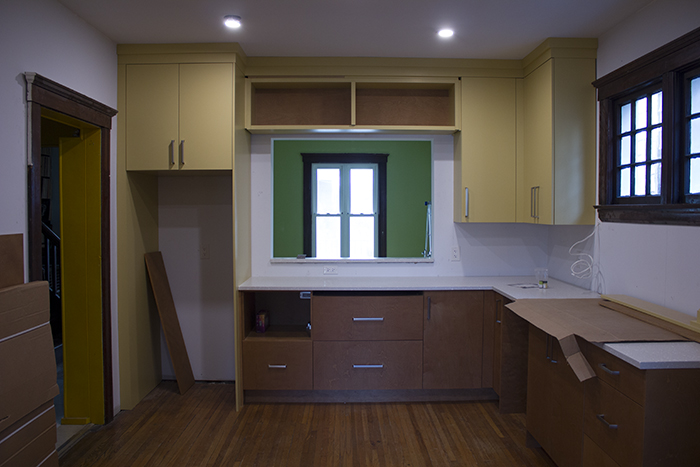 An unfinished kitchen with yellow and wood tone cabinets, hardwood floor, white wall, and a window opening into a room beyond which is painted green and has a tall window in the centre.