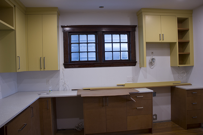 An unfinished kitchen with yellow and wood tone cabinets and white walls with an older style window trimmed with dark wood in the centre.