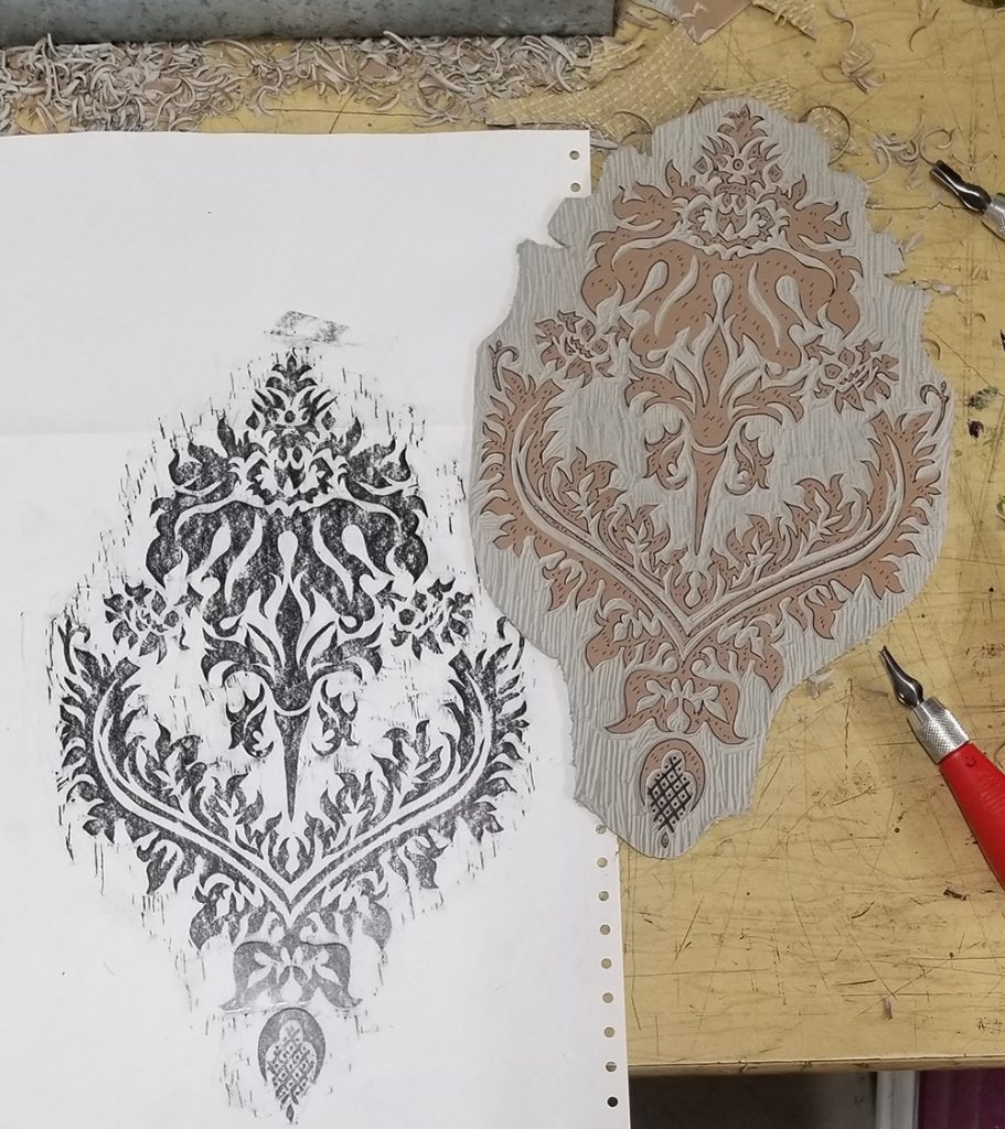 a carved linoleum block sitting next to a graphite rubbing of the block on paper, with carving tools scattered around