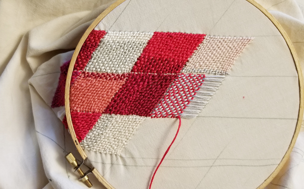 white fabric in an embroidery hoop with woven stitched diamonds in shades of red