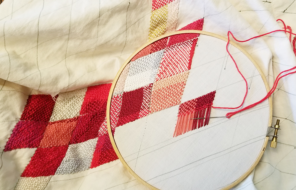 white fabric in an embroidery hoop with woven stitched diamonds in shades of red