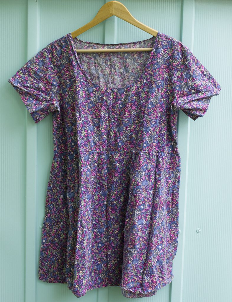 a loose cotton dress with short sleeves, in a small blue and pink floral print, hanging against a seafoam green steel garden shed