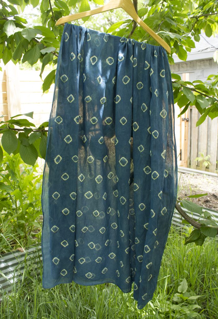indigo dyed cloth with an allover pattern of tiny yellow diamonds, hanging from a cherry tree in an overgrown garden